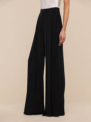 Wearing the Nova Pleated Wide-Leg Pant in black with a white sleeveless top, a person stands against a beige background. The photo is cropped at the shoulders.