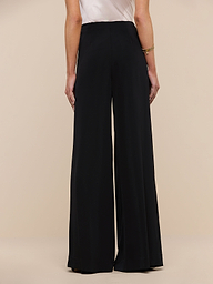 A person stands facing away against a plain, light background, showing only their lower back and legs as they wear the Nova Pleated Wide-Leg Pant in black with a tailored, high-waisted fit beneath a white sleeveless top.