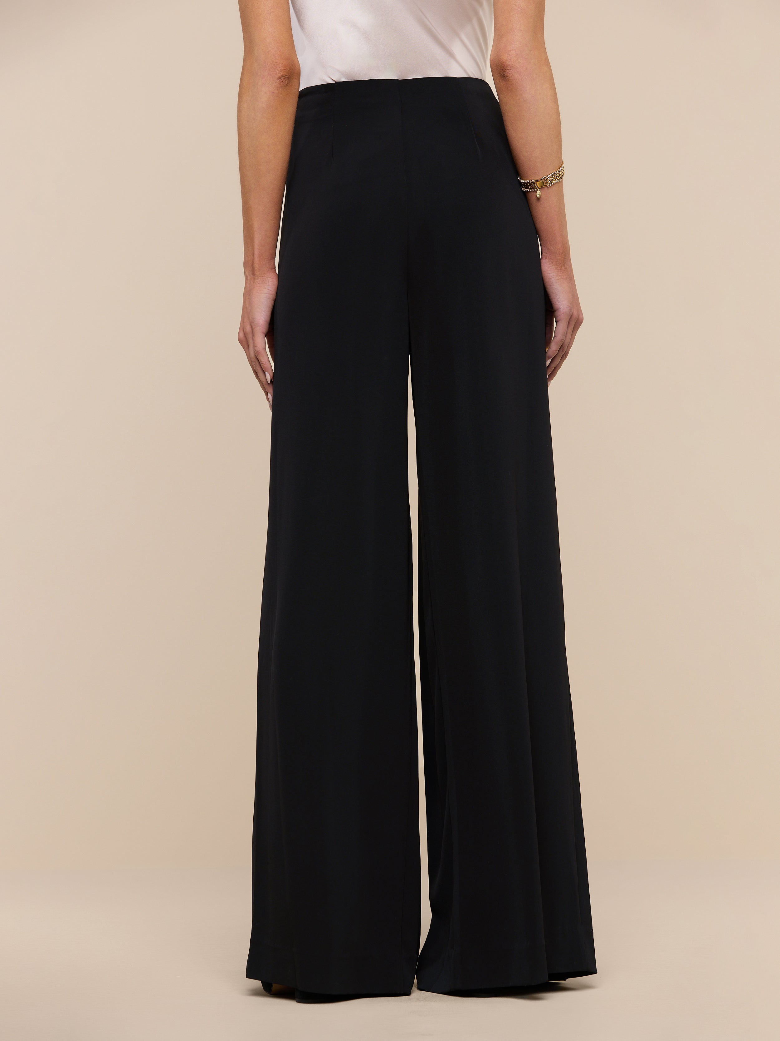 A person stands facing away against a plain, light background, showing only their lower back and legs as they wear the Nova Pleated Wide-Leg Pant in black with a tailored, high-waisted fit beneath a white sleeveless top.