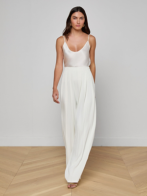 A woman stands indoors on a wooden floor, wearing the Nova Pleated Wide-Leg Pant with a sleeveless white top. She has long brown hair, a neutral expression, and looks directly at the camera.