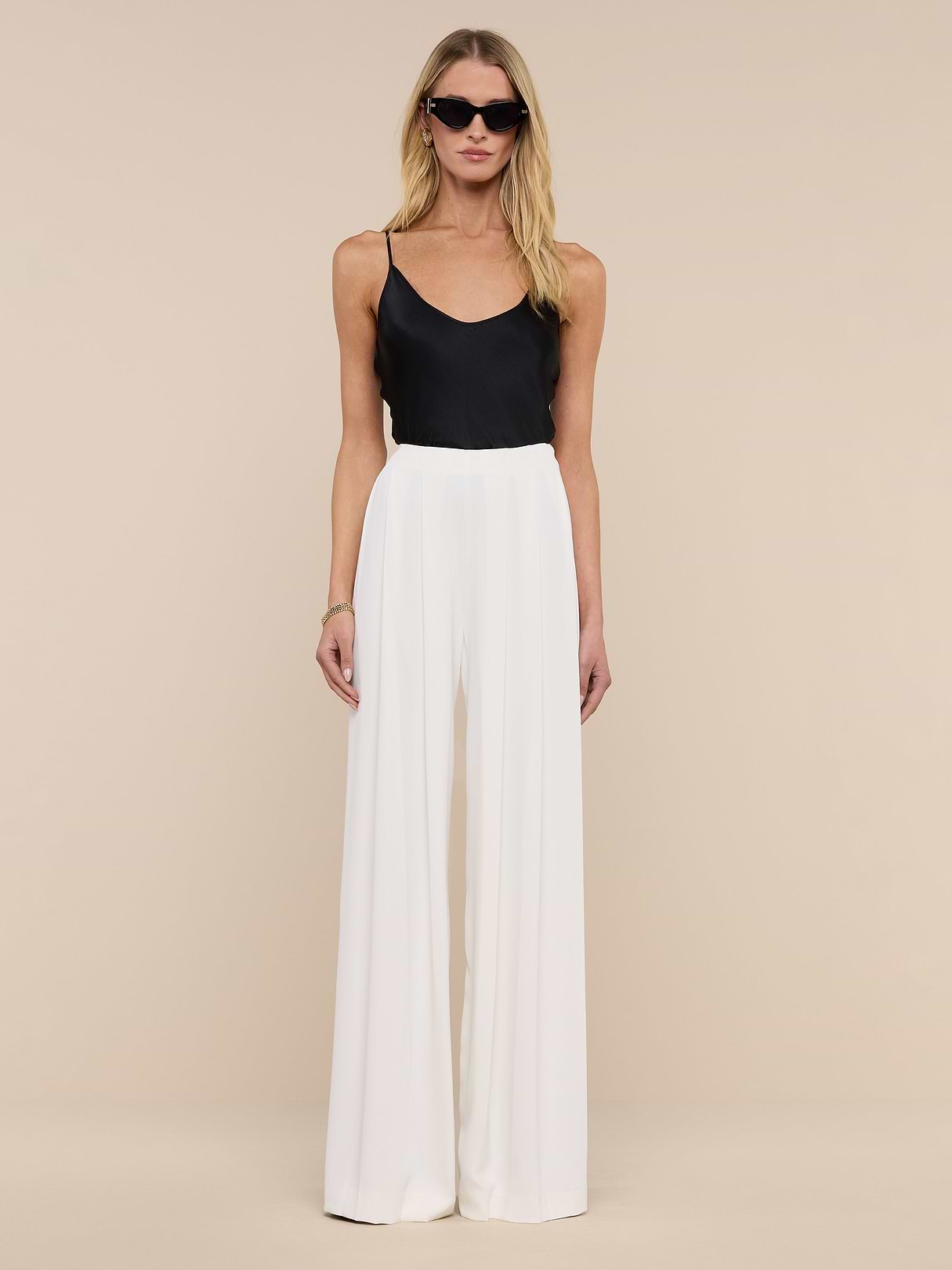 A woman in sunglasses wears a black sleeveless top and the Nova Pleated Wide-Leg Pant in ivory, standing against a plain white background.