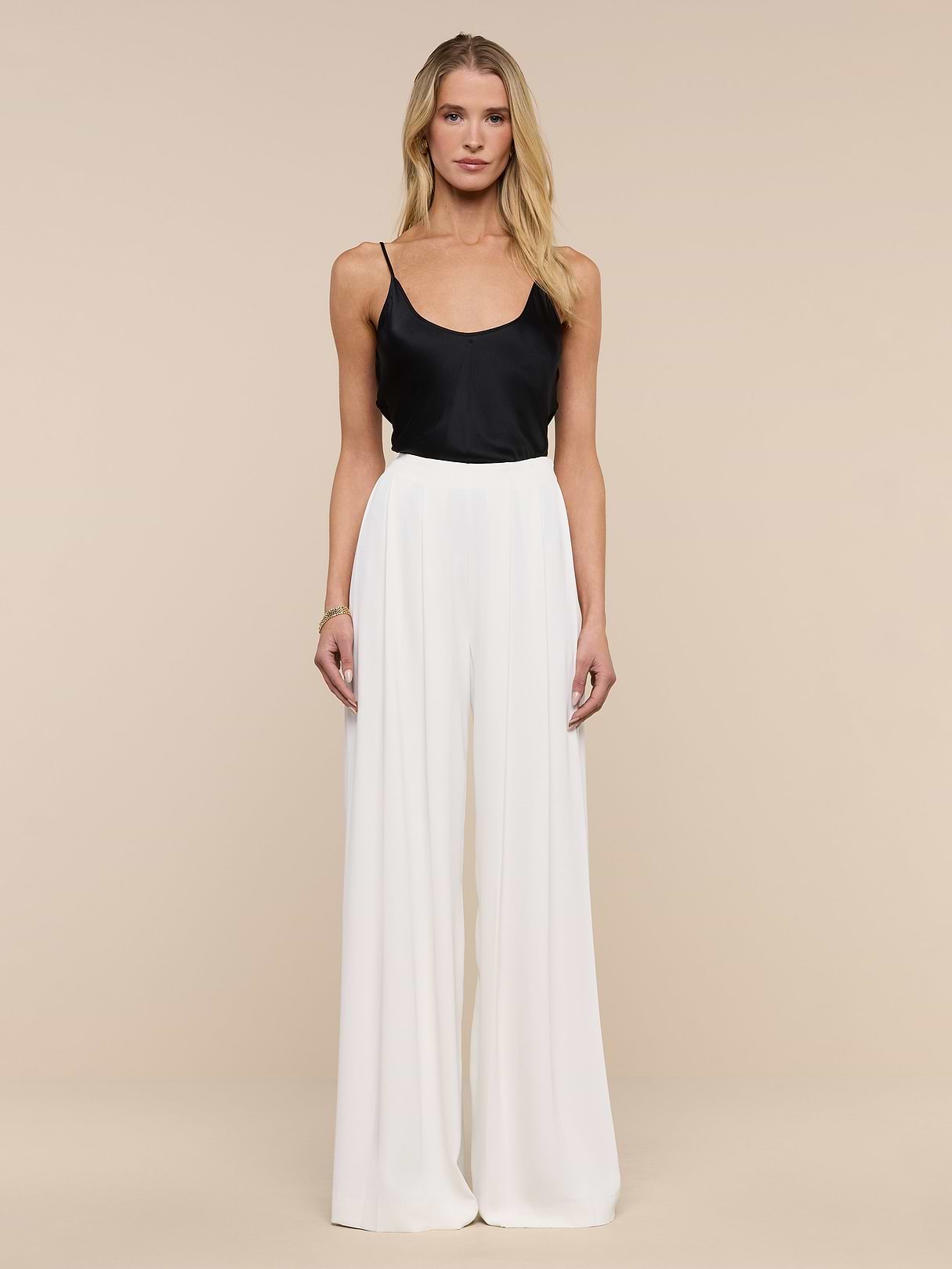 A woman stands facing forward against a plain white background, wearing a black sleeveless top paired with the Nova Pleated Wide-Leg Pant in ivory.