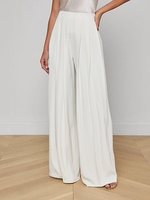 A person wears the Nova Pleated Wide-Leg Pant with a tucked-in sleeveless top, standing on a light wood floor, one hand on their hip. Their head is not visible in the image.