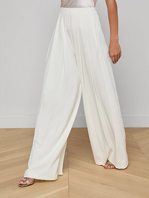 Someone stands on a wooden floor against a plain light-colored wall, wearing the Nova Pleated Wide-Leg Pant in suiting fabric paired with metallic open-toe heels.