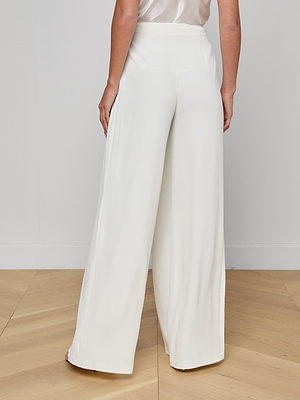 A person stands on a light wooden floor, shown from the back, wearing the Nova Pleated Wide-Leg Pant in white suiting fabric with a loose, high-rise fit and floor-grazing wide legs, styled with a light satin top.