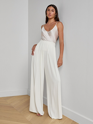 A woman stands against a white wall wearing a sleeveless satin top and the Nova Pleated Wide-Leg Pant in suiting fabric, creating a chic minimalist look as she poses on a light wood floor.
