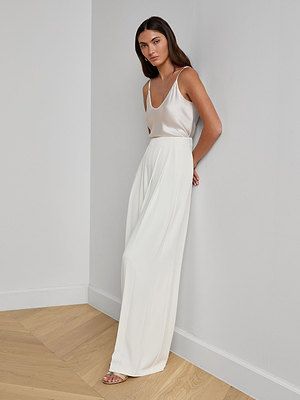 A woman with long brown hair stands against a white wall, wearing a sleeveless white top and the Nova Pleated Wide-Leg Pant with beige heeled sandals. The setting features light wood flooring.