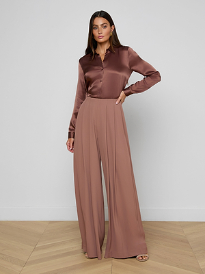 A woman with long straight hair and a neutral expression stands indoors against a light wall, wearing the Nova Pleated Wide-Leg Pant in fluid suiting fabric, paired with a silky brown long-sleeve blouse and heeled sandals.