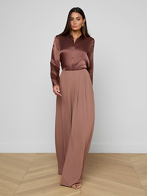 A woman stands on a light wooden floor against a plain light gray wall, wearing the Nova Pleated Wide-Leg Pant with a shiny brown button-up blouse and open-toed heels.