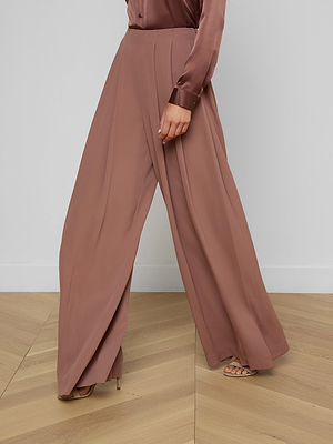 A person stands on a wooden floor, showing only their lower body. They're wearing the Nova Pleated Wide-Leg Pant in mauve fluid suiting fabric with a matching silk blouse and elegant heeled sandals.