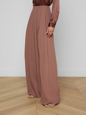 A person wears the Nova Pleated Wide-Leg Pant in mauve, made from a fluid suiting fabric, paired with a long-sleeved satin blouse. Only their lower body is shown as they stand on a light wooden floor against a plain white wall.