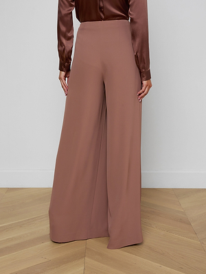 A person stands facing away, wearing the Nova Pleated Wide-Leg Pant in mauve fluid suiting fabric with a matching long sleeve satin blouse, set against a light wall and chevron wood floor.