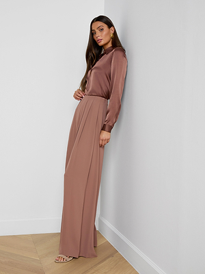 A woman with long hair and gold earrings stands against a white wall wearing the Nova Pleated Wide-Leg Pant in fluid suiting fabric, paired with a silky mauve blouse and open-toed heels.