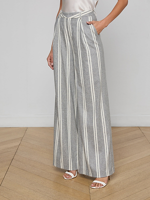 Wearing the Allanah Striped Trouser—high-waisted wide-leg gray pants in an Italian wool blend with white stripes—paired with a light sleeveless top and white heeled sandals, this person stands on a wood floor.