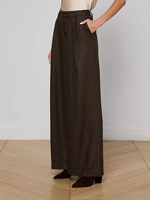 A person stands with one hand in their pocket, wearing L'AGENCE Allanah Pinstriped Trousers—high-waisted, wide-leg, dark brown pinstripes in recycled wool-blend—paired with brown pointed-toe shoes on a wooden floor.