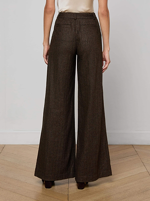 A person stands with their back to the camera on a wooden floor, wearing L'AGENCE Allanah Pinstriped Trousers in recycled wool-blend, featuring a high-waisted, wide-leg brown modern design against a plain white wall.