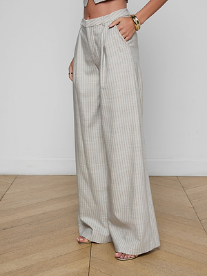 A person wears the Allanah Pinstriped Trouser—high-waisted, wide-leg, light beige with thin vertical stripes—in Italian suit fabric, styled with a cropped top, gold bangles, and open-toe heels on a wooden floor before a white wall.