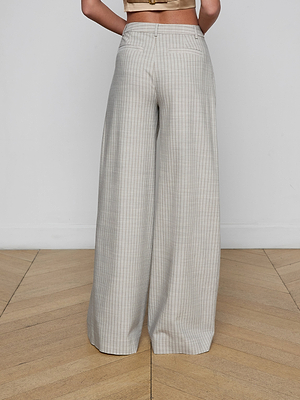 A person stands facing away, wearing the Allanah Pinstriped Trouser—high-waisted, wide-leg modern pants crafted from Italian suit fabric in a light plaid. The beige cropped top complements the wooden floor and plain white wall.