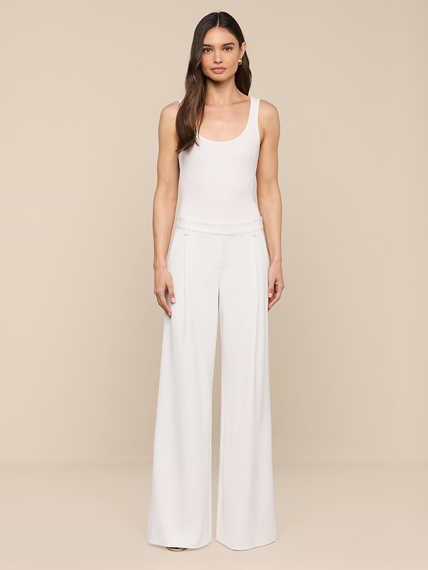 A woman with long brown hair and a neutral expression stands against a plain white background, wearing modern, wide-leg Allanah Trousers crafted from stretch suit fabric for a flattering fit, paired with a sleeveless white top.