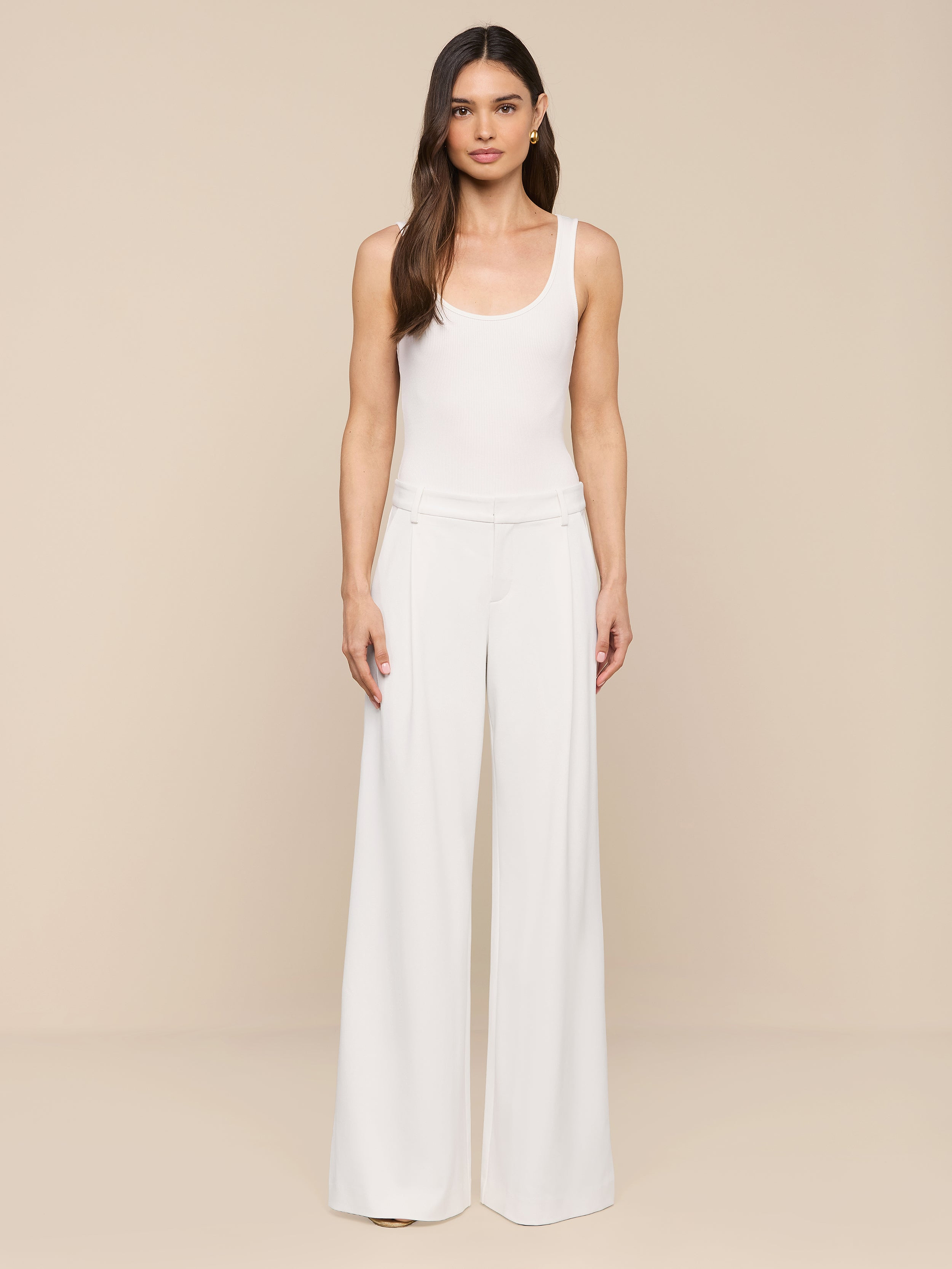 A woman with long brown hair and a neutral expression stands against a plain white background, wearing modern, wide-leg Allanah Trousers crafted from stretch suit fabric for a flattering fit, paired with a sleeveless white top.