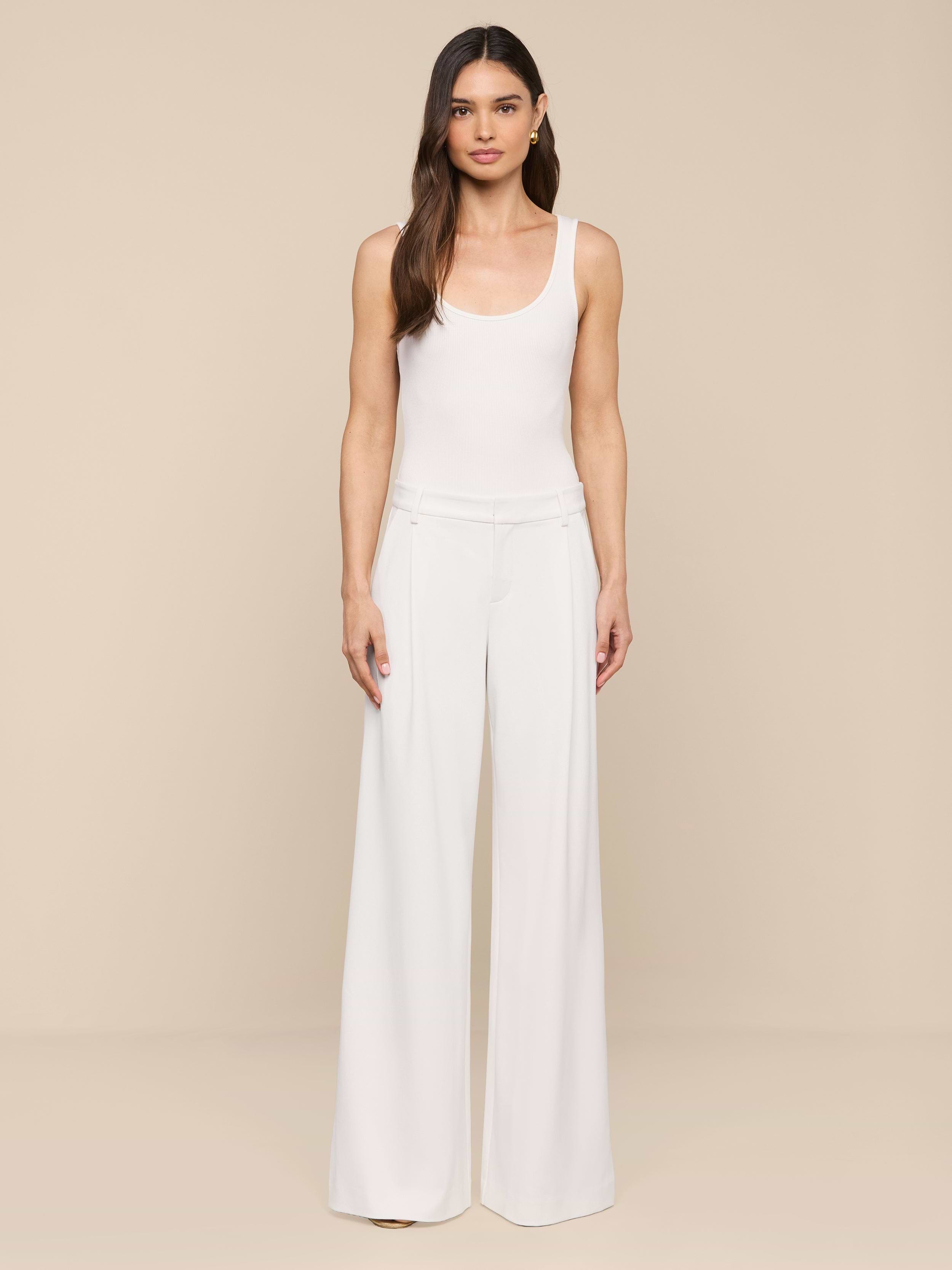 A woman with long brown hair and a neutral expression stands against a plain white background, wearing modern, wide-leg Allanah Trousers crafted from stretch suit fabric for a flattering fit, paired with a sleeveless white top.