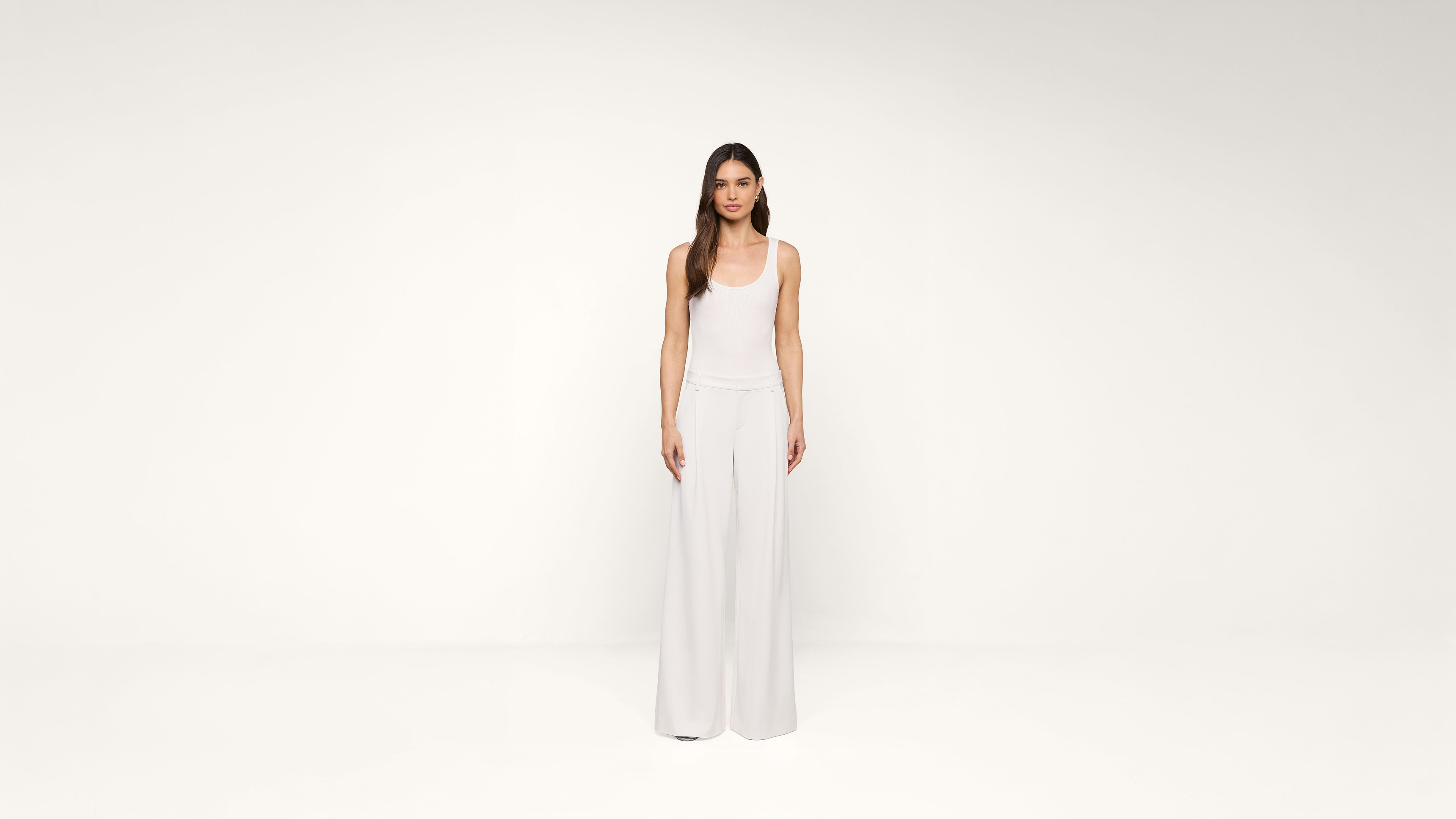 A woman with long hair stands facing forward, wearing a white sleeveless top and wide-legged white pants, against a plain white background.