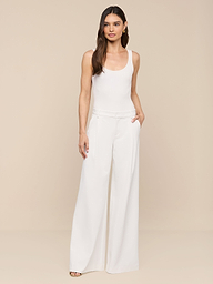A woman with long brown hair stands against a plain white background, wearing a white sleeveless top paired with the Allanah Trouser—high-waisted, wide-leg pants designed in a modern and flattering fit. She has one hand in her pocket and is looking at the camera.