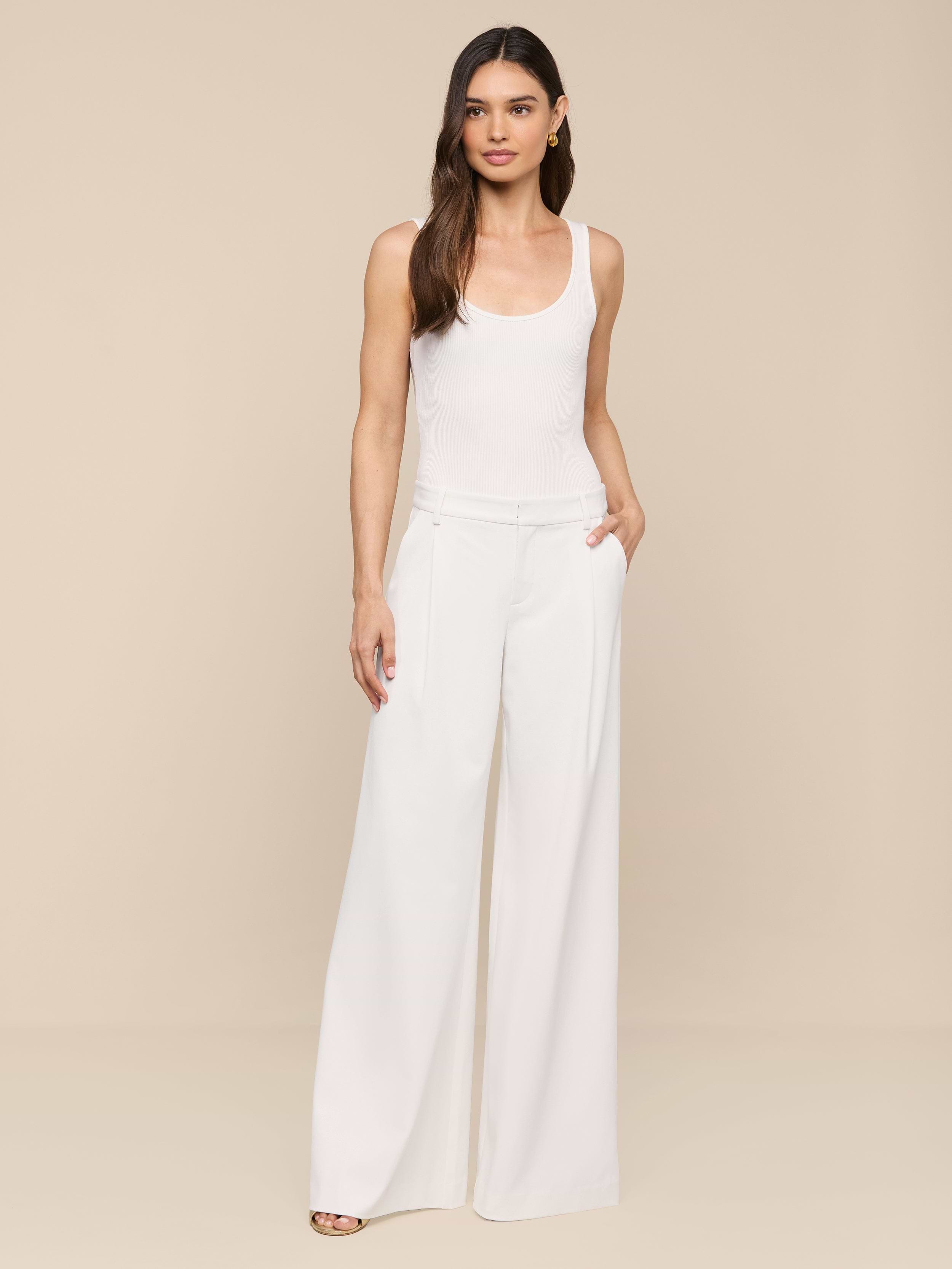A woman with long brown hair stands against a plain white background, wearing a white sleeveless top paired with the Allanah Trouser—high-waisted, wide-leg pants designed in a modern and flattering fit. She has one hand in her pocket and is looking at the camera.