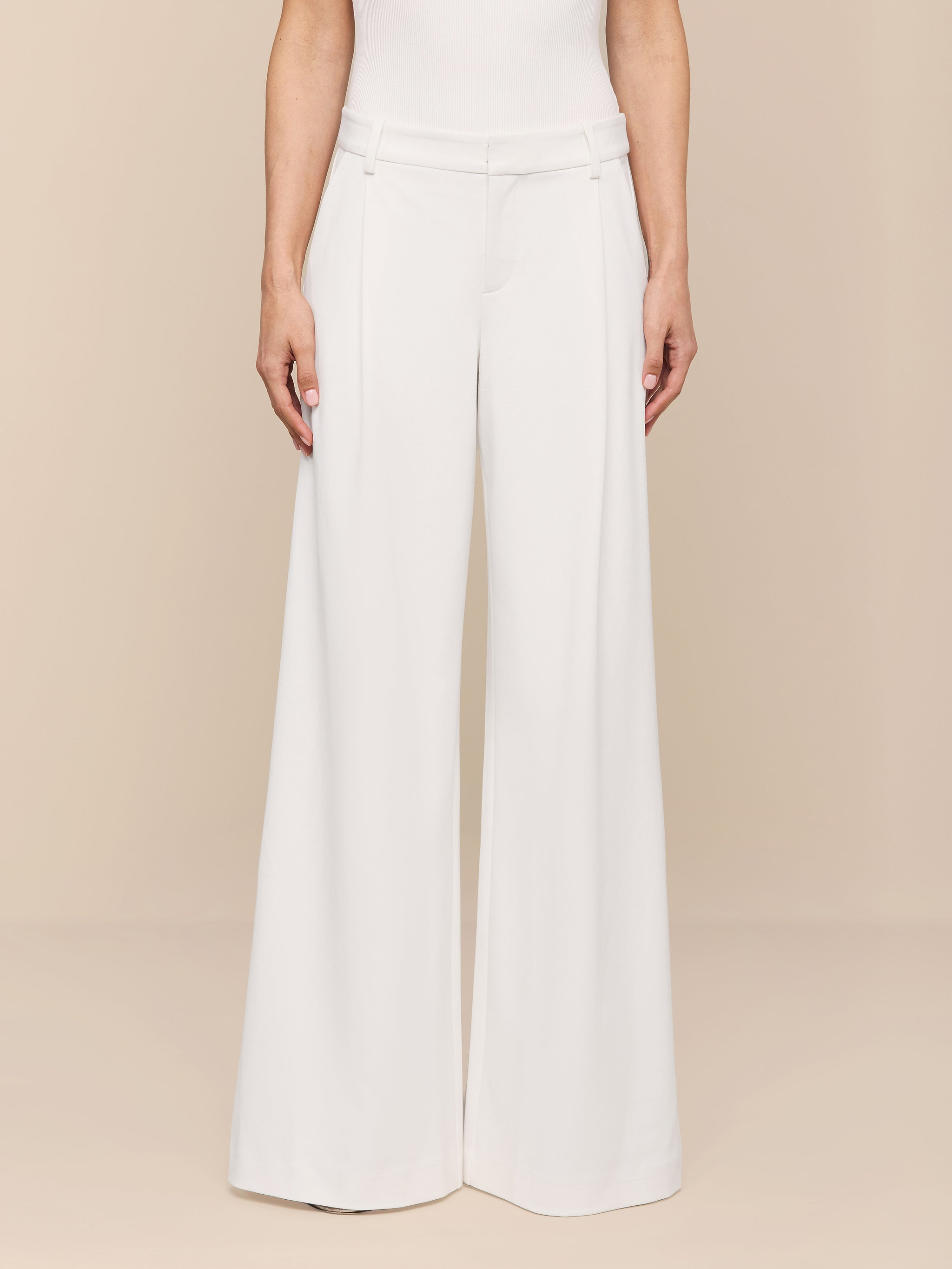 A person stands against a plain white background, shown from the shoulders down, wearing the Allanah Trouser—high-waisted, wide-leg trousers in white stretch suit fabric—paired with a sleeveless white top.
