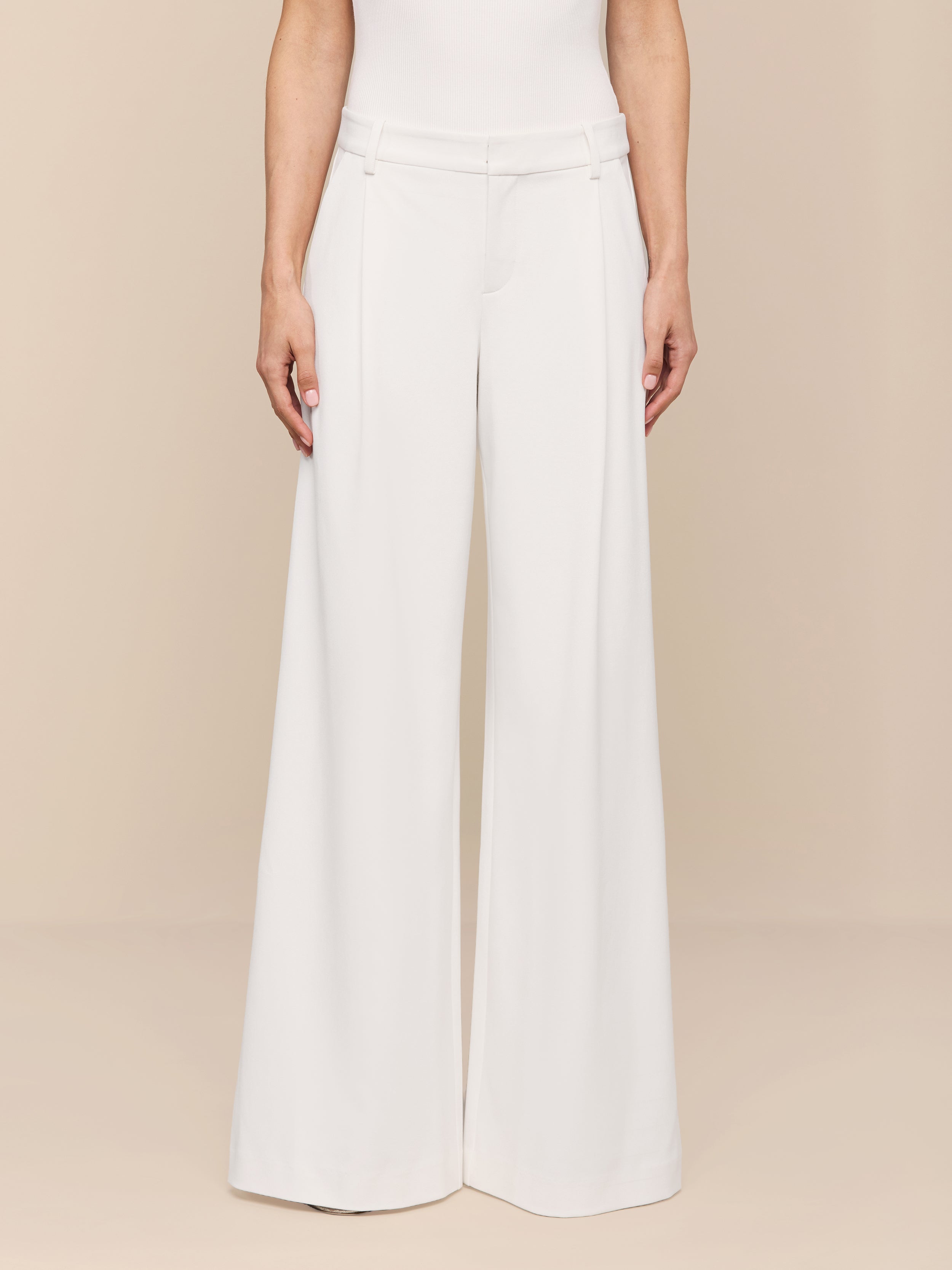 A person stands against a plain white background, shown from the shoulders down, wearing the Allanah Trouser—high-waisted, wide-leg trousers in white stretch suit fabric—paired with a sleeveless white top.
