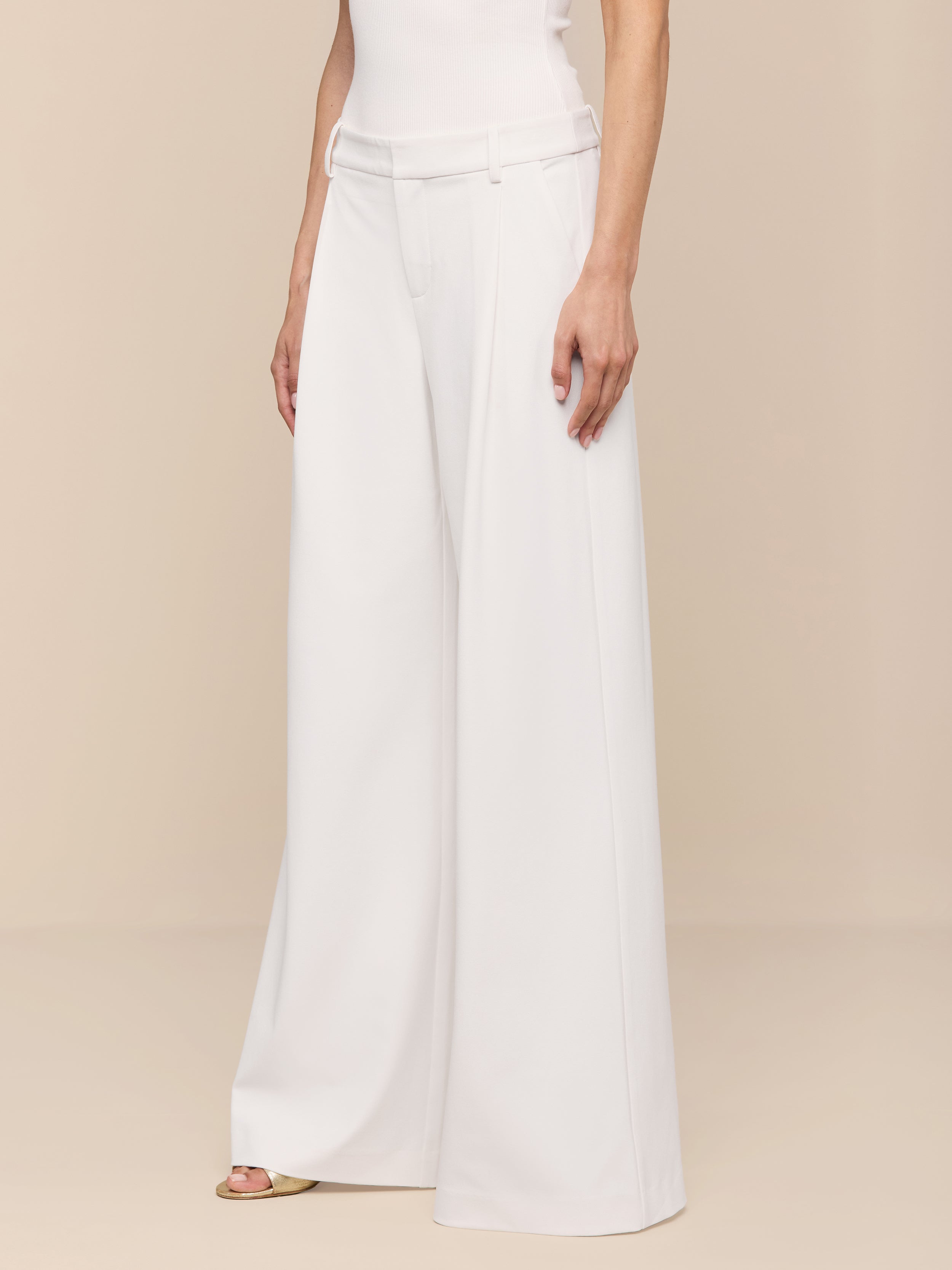 A person stands against a plain white background, showcasing only the lower half of their body in high-waisted, wide-leg Allanah Trousers crafted from stretch suit fabric. The modern design and flattering fit of these white trousers are highlighted, paired with a sleeveless white top.