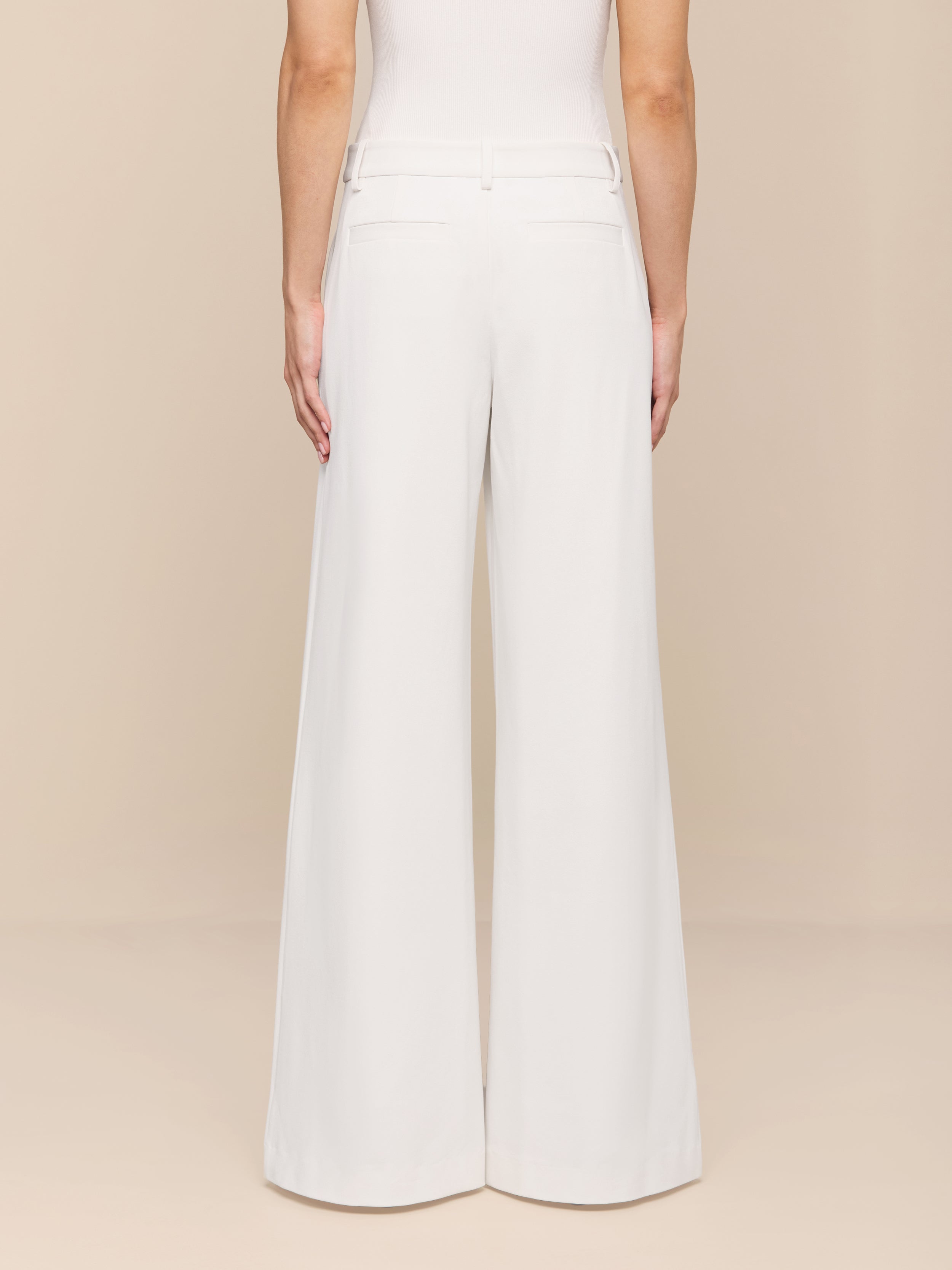 A person is shown from the back against a plain white background, wearing the Allanah Trouser—high-waisted, wide-leg white trousers crafted from modern trouser stretch suit fabric—paired with a sleeveless white top for a flattering fit.
