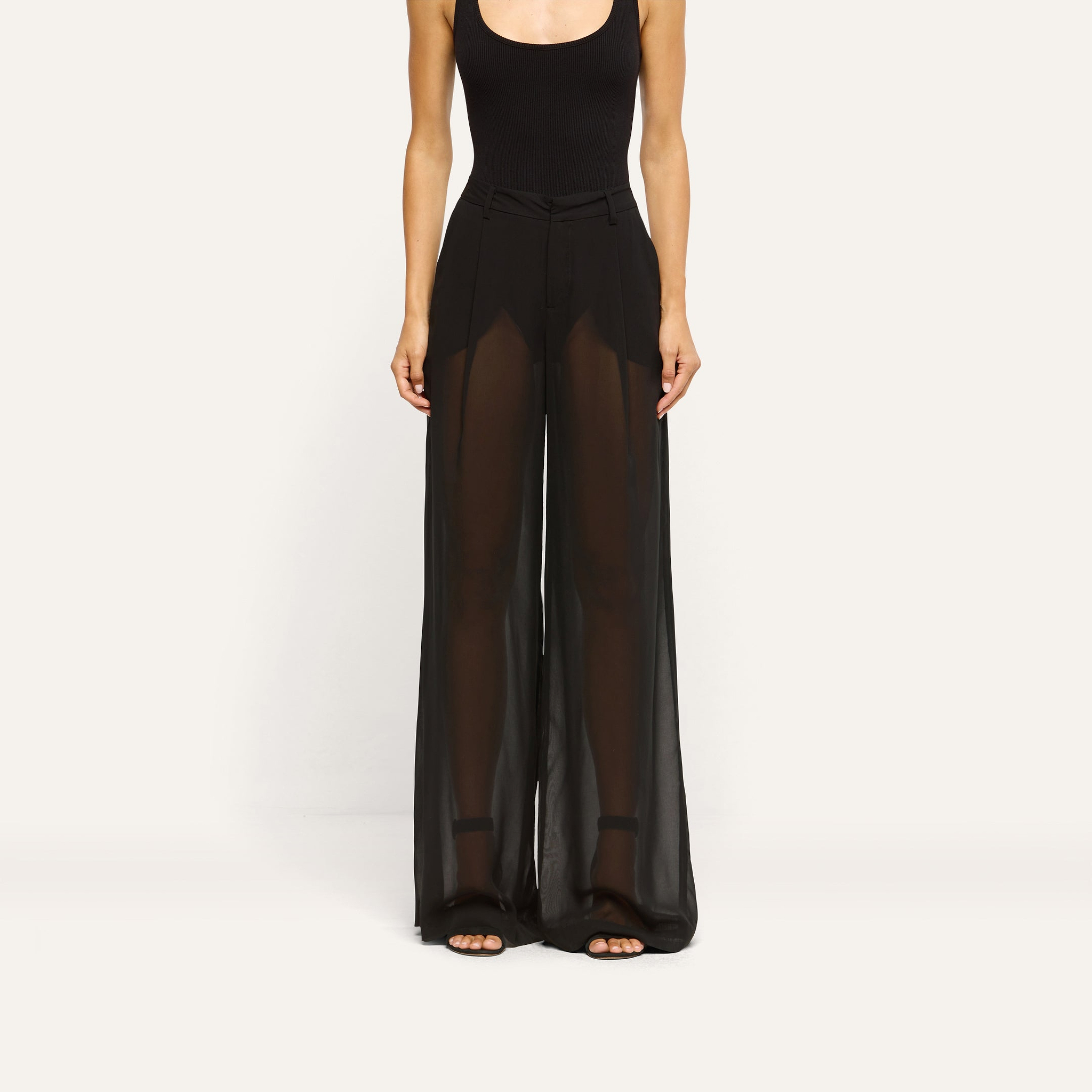 A person wearing a black bodysuit and Allanah Sheer Trouser, featuring mid-rise, sheer chiffon fabric with a floor-length, wide-leg silhouette.