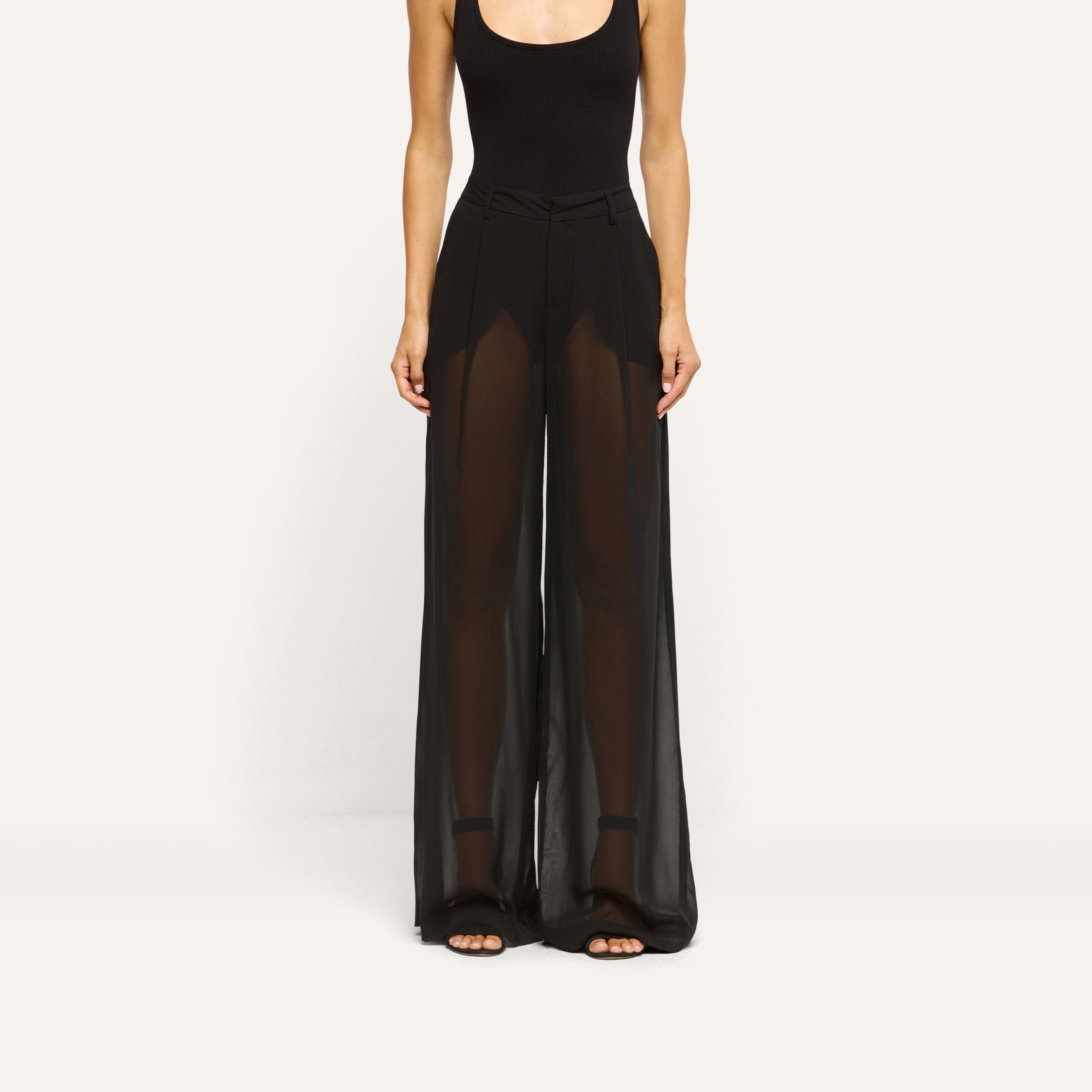 A person wearing a black bodysuit and Allanah Sheer Trouser, featuring mid-rise, sheer chiffon fabric with a floor-length, wide-leg silhouette.