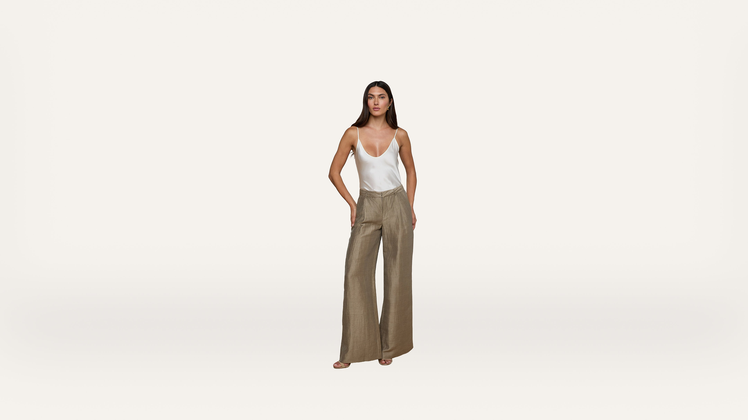 A woman stands against a plain white background wearing a white tank top and high-waisted, wide-leg beige trousers. She has long hair and a neutral expression, with her hands in her pockets.