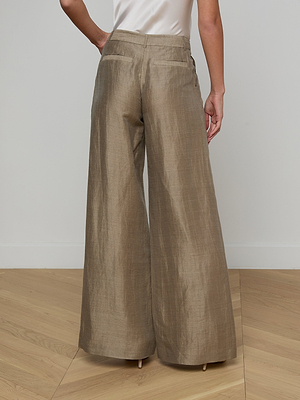 A person stands on a wooden floor against a plain light wall, wearing the Allanah Linen-Blend Trouser—wide-legged, high-waisted, and light brown with a modern, flattering fit.