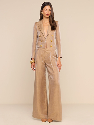 A woman stands against a beige background, wearing the Allanah Linen-Blend Trouser—a shimmery beige pantsuit with gold buttons and modern wide-leg linen-blend pants—paired with nude heels and gold bracelets. Her long, wavy brown hair frames her neutral expression.