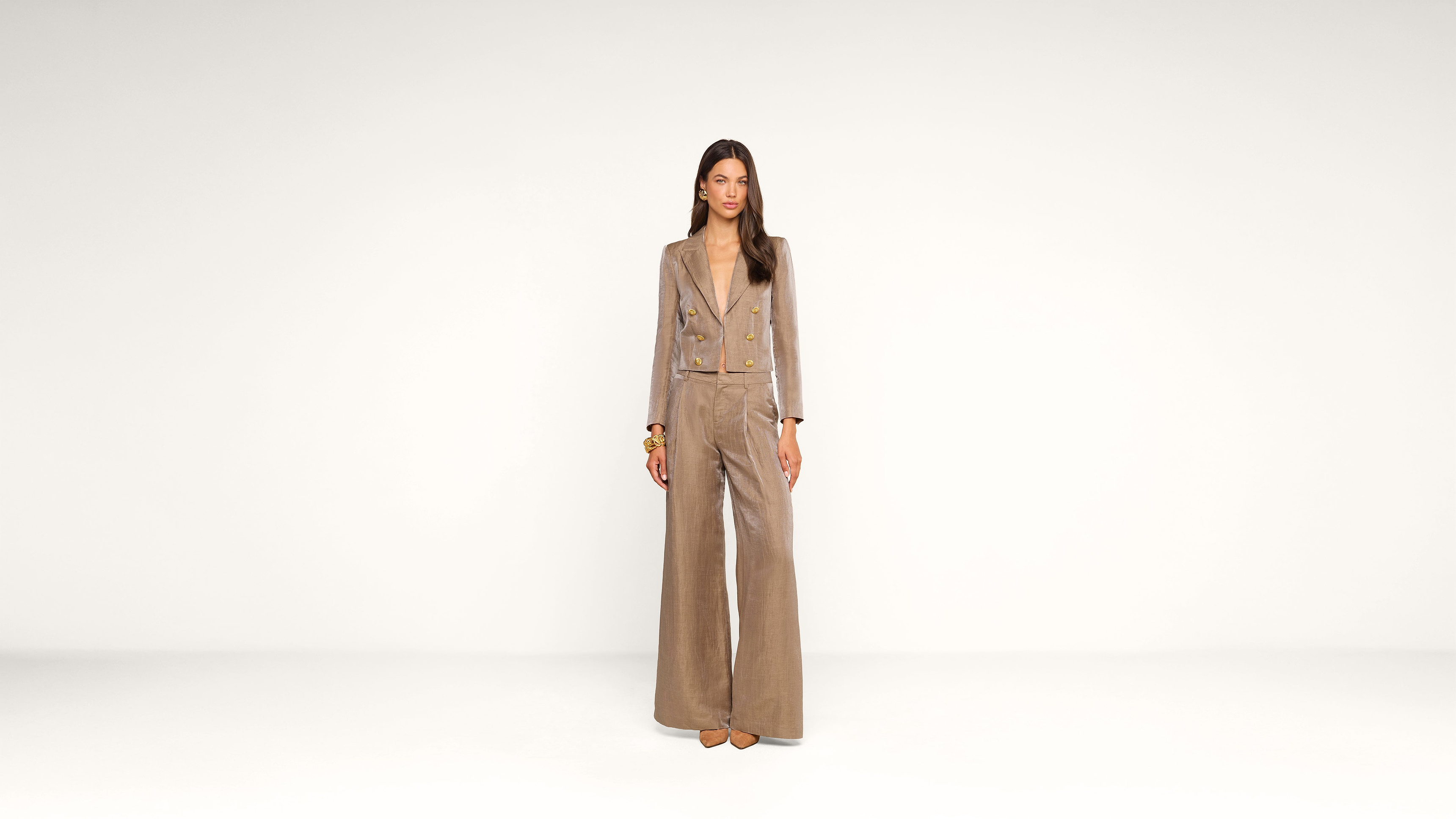A woman stands against a plain white background, wearing a tan wide-leg pantsuit with a matching blazer and brown shoes. She has long straight hair and a neutral expression, with her arms relaxed at her sides.
