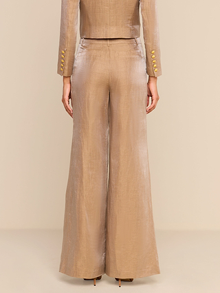 A person stands facing away, wearing shiny, latte brown Allanah Linen-Blend Trousers and a matching jacket with gold buttons. The modern trouser silhouette and tailored jacket create an elegant look against the neutral background.