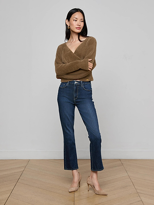 A woman with long dark hair stands on a wooden floor against a light gray wall, wearing L’AGENCE’s Mira Cropped Bootcut Jeans in stretch denim, a brown sweater, and beige heels. Her arms are crossed as she looks to the side.