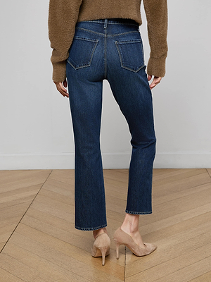 A person stands on a wood floor, facing away from the camera, wearing a brown sweater, L'AGENCE Mira Cropped Bootcut Jean in dark blue stretch denim, and beige high-heeled shoes.