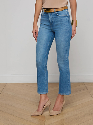 A person stands on a wooden floor, wearing the Mira Cropped Bootcut Jean in light blue with a tan belt, beige top, and tan heels. The photo is cropped from the shoulders down.