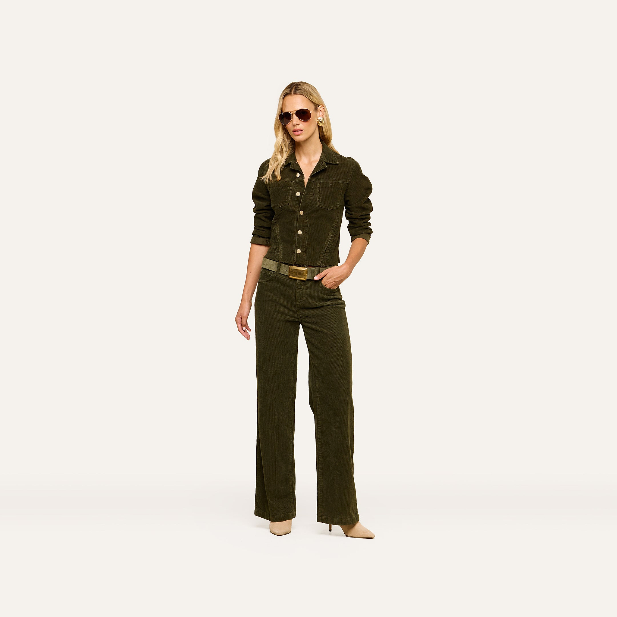 A woman wearing sunglasses, a dark green corduroy jacket, the Scottie Corduroy Pant in a matching high-rise wide-leg silhouette, a belt, and beige heels stands against a plain light background, capturing the essence of fall 2026 fashion.