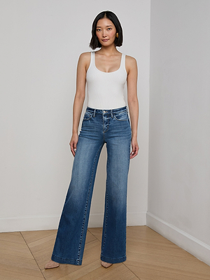 A woman with short dark hair stands indoors on a wooden floor, wearing a white sleeveless top and the Scottie Wide-Leg Jean in high-rise blue stretch denim, looking at the camera with a neutral expression.