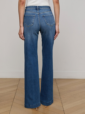 A woman faces away from the camera, wearing Scottie Wide-Leg Jeans in blue stretch denim with back pockets and a white sleeveless top. The wide-leg jeans reach her shoes as she stands on a wooden floor.