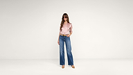 A woman stands in a minimal studio, wearing sunglasses, a silky pink blouse, blue wide-leg jeans, and tan heels. She has long hair and one hand in her pocket, posing confidently against a plain background.