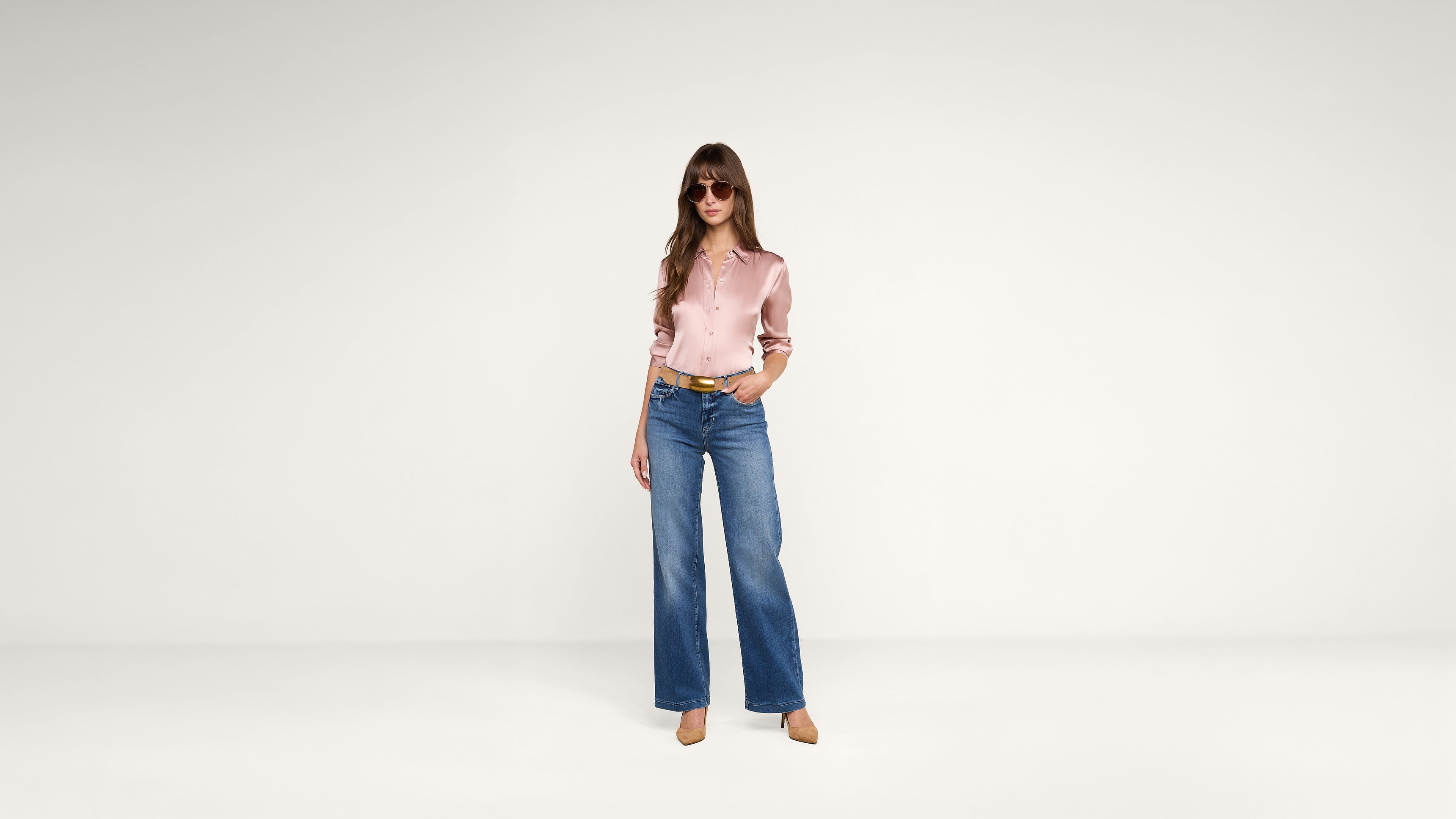 A woman stands in a minimal studio, wearing sunglasses, a silky pink blouse, blue wide-leg jeans, and tan heels. She has long hair and one hand in her pocket, posing confidently against a plain background.