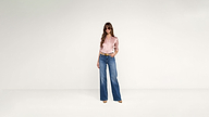 A woman in a stylish pink button-down shirt, wide-leg blue jeans, and a gold belt stands against a white background wearing sunglasses.