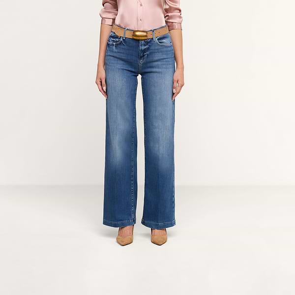 Una persona che indossa dei jeans Scottie Wide-Leg con una cintura color cuoio, una camicetta di raso rosa chiaro e scarpe con tacco a punta color cuoio è in piedi davanti a uno sfondo beige a tinta unita.