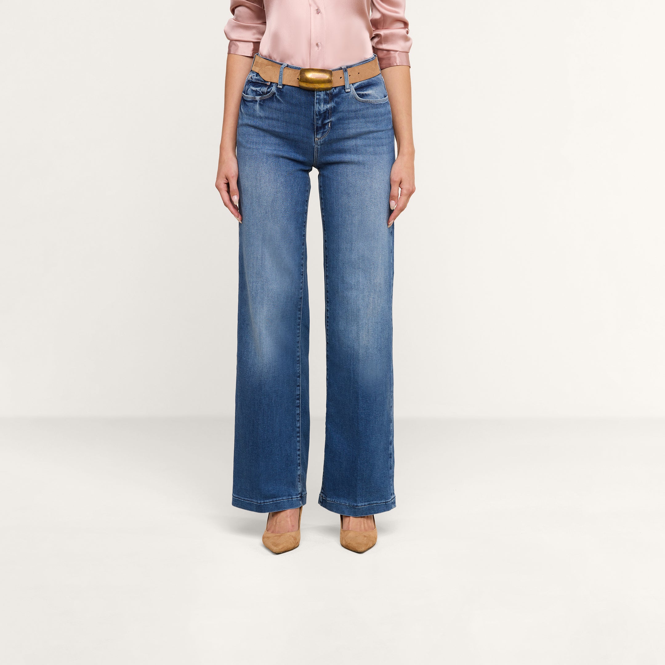Una persona che indossa dei jeans Scottie Wide-Leg con una cintura color cuoio, una camicetta di raso rosa chiaro e scarpe con tacco a punta color cuoio è in piedi davanti a uno sfondo beige a tinta unita.